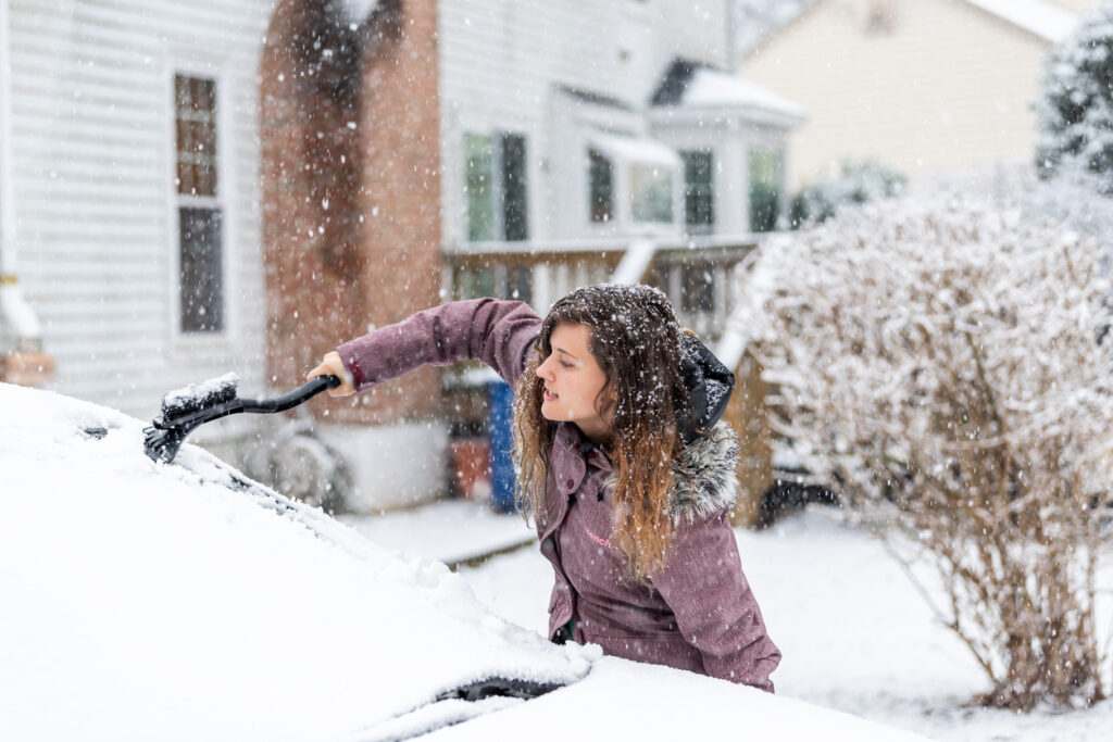 Woman is freezing while cleaning car's windshield from snow and ice. Snowy winter day.