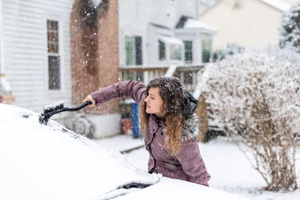 Woman is freezing while cleaning car's windshield from snow and ice. Snowy winter day.