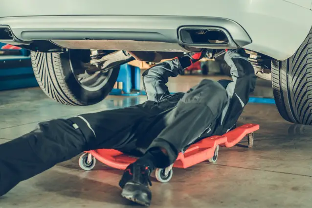 Car mechanic is fixing old car's suspension, he is laying on the trolley 