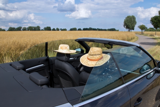 Nice empty convertible car on the countryside road besides yellow field. Two hats on each car seat. Summer outside.